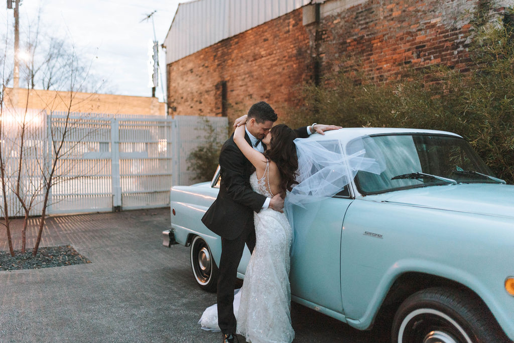 bride and groom posing in front of a blue car